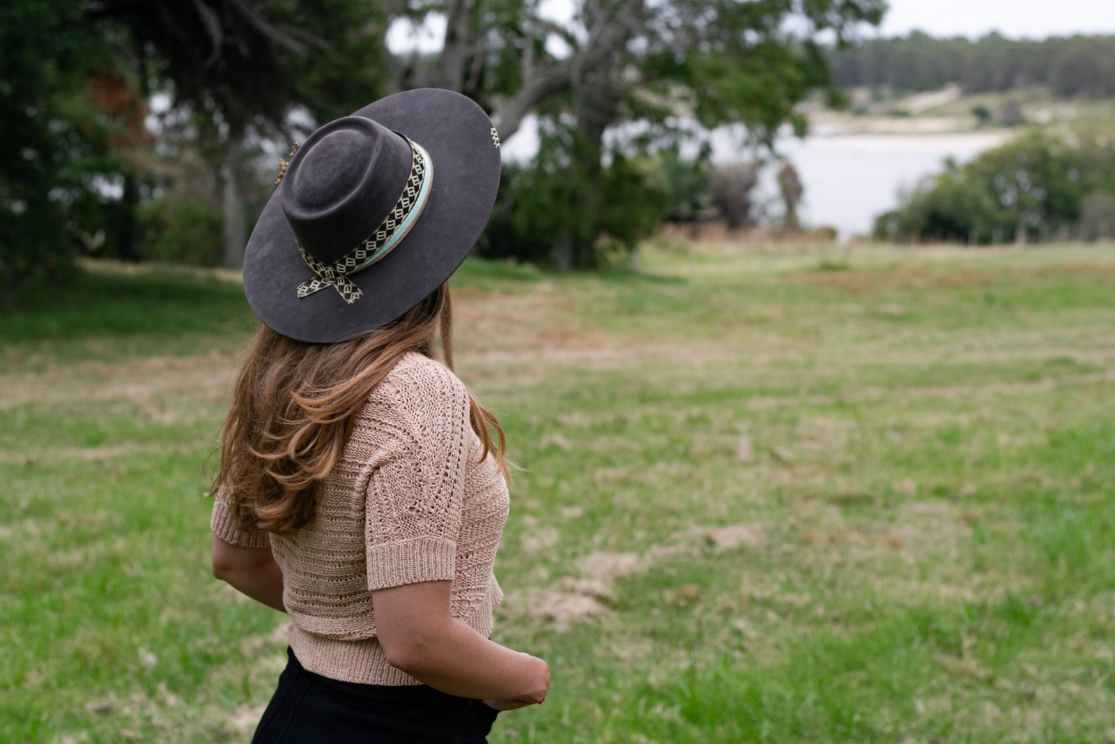 Woman wearing a black wide-brim felt hat standing in a grassy field near a lake